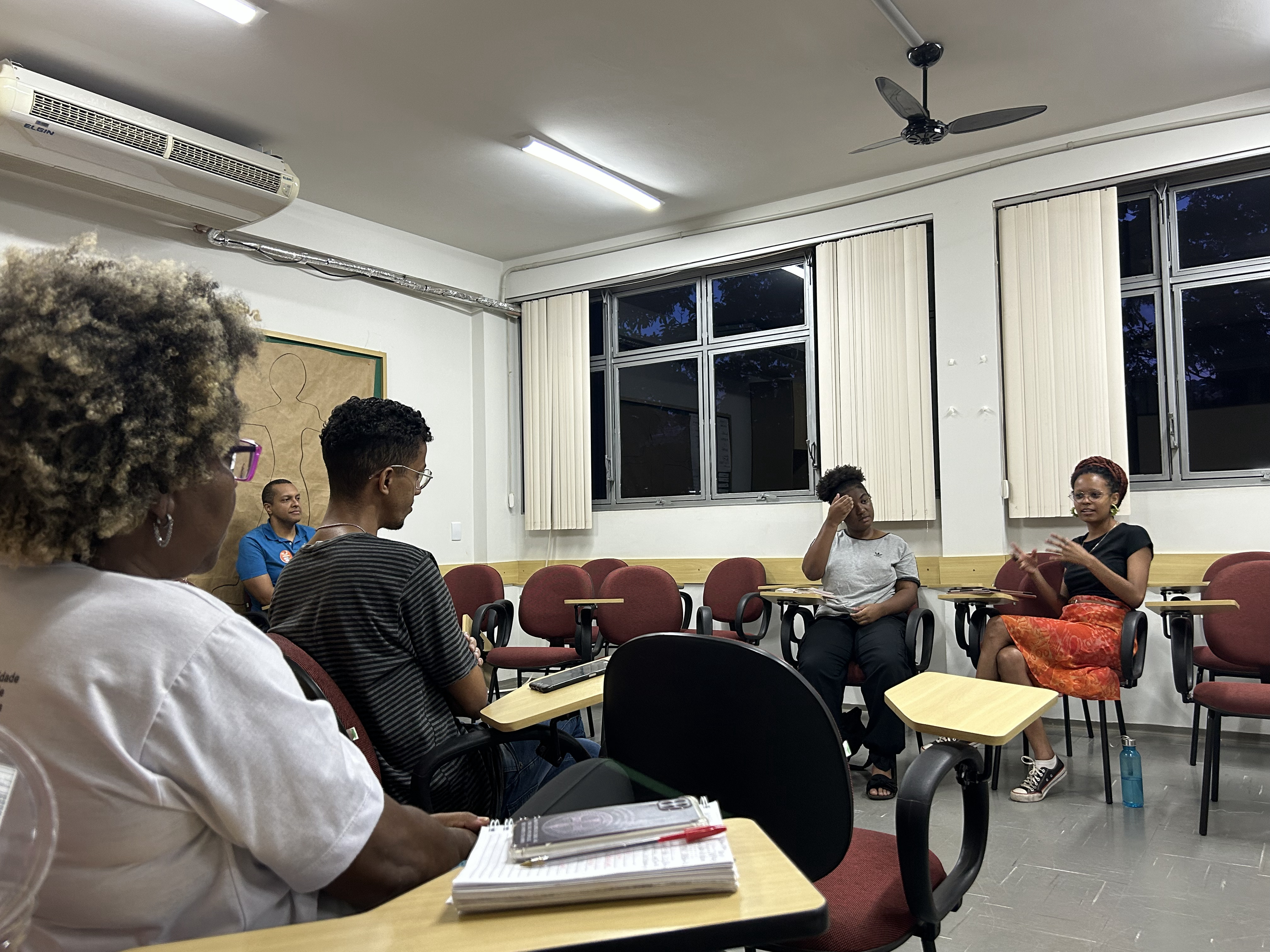 Foto colorida com grupo de pessoas negras em semicírculo, em sala de estudos, debatendo texto