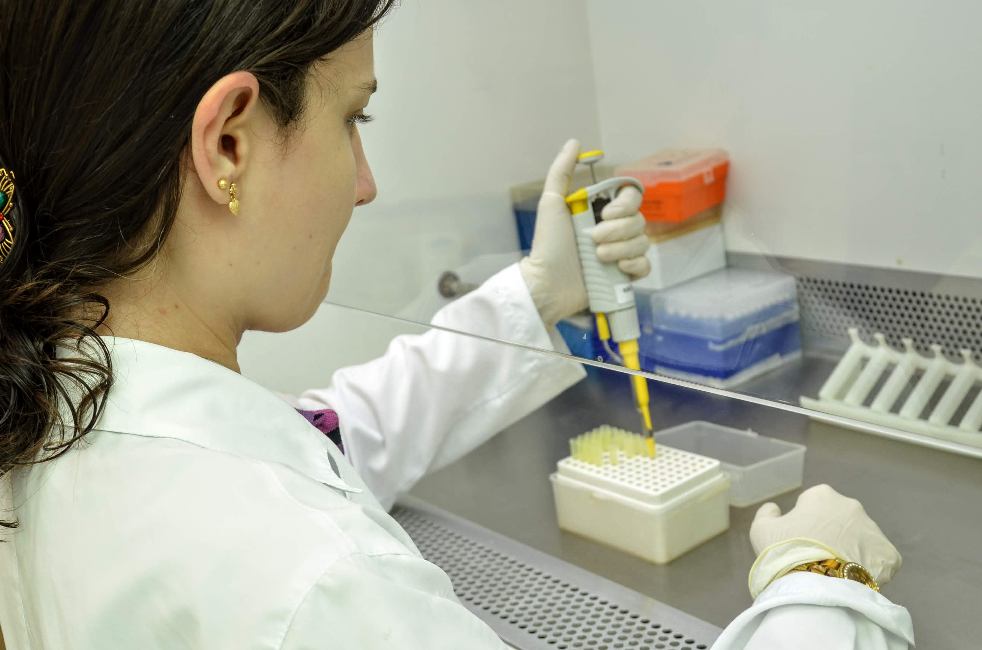 Foto colorida com mulher de costas vestida com jaleco branco, trabalhando em laboratório