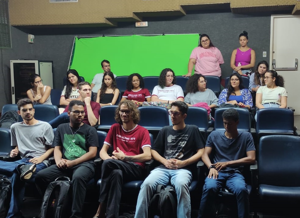 Na imagem, os estudantes estão sentados em um auditório com o fundo verde e com duas servidoras da Dirco em pé, ao fundo