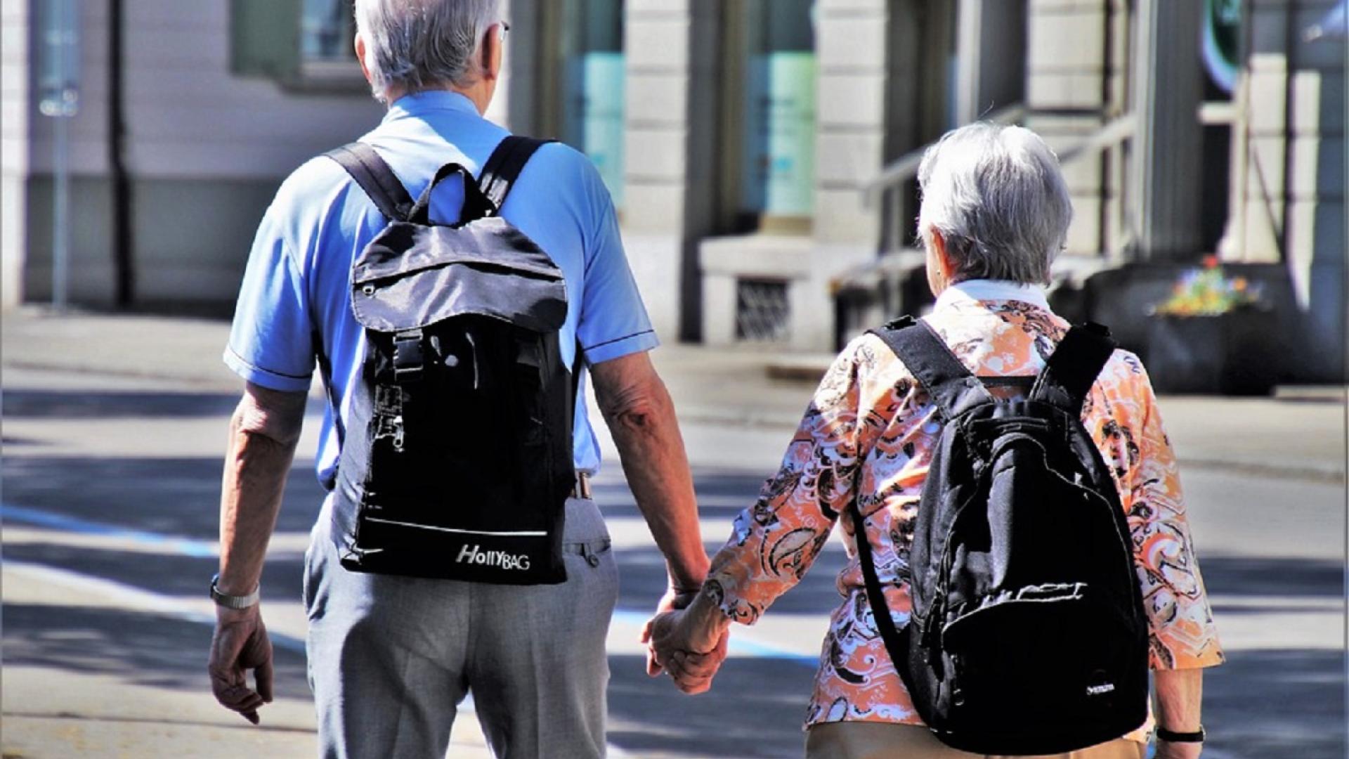 Imagem de um homem e uma mulher de costas, ambos com cabelos grisalhos e mochilas na costas, caminhando de mãos dadas