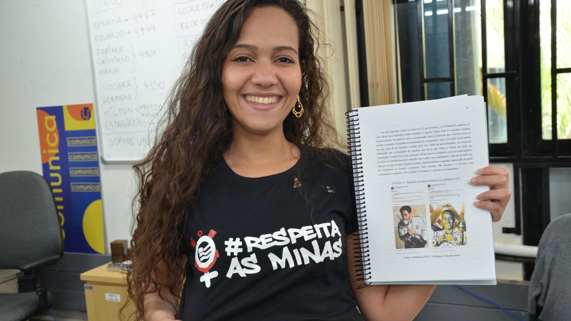 A autora da pesquisa, Ítana Santos, segurando uma versão impressa da monografia e vestida com uma camisa com o nome do trabalho: "Respeita as minas"