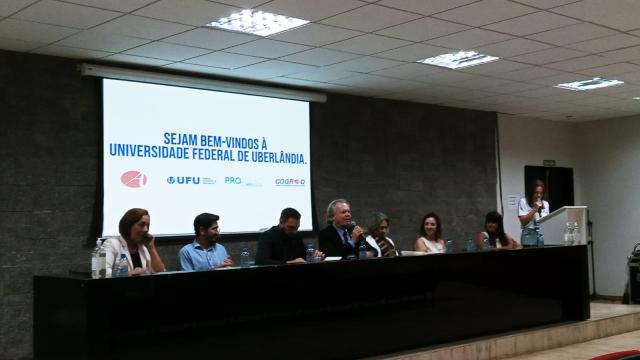 Imagem mostra ao fundo um telão digital escrito 'Sejam bem-vindos à Universidade Federal de Uberlândia'; à frente, estão sete pessoas sentadas diante de uma mesa e uma em pé segurando um microfone