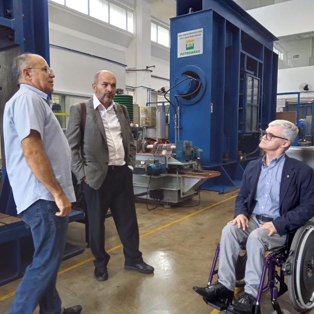 Três homens, sendo um deles cadeirante, conversando em um laboratório de engenharia mecânica
